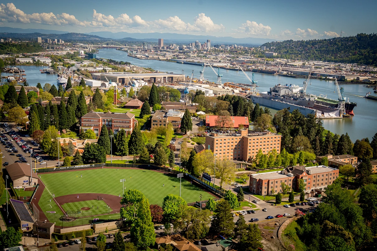 A scenic aerial view of a riverside city featuring a college campus with a baseball field in the foreground, surrounded by trees, buildings, and a shipyard along the river.